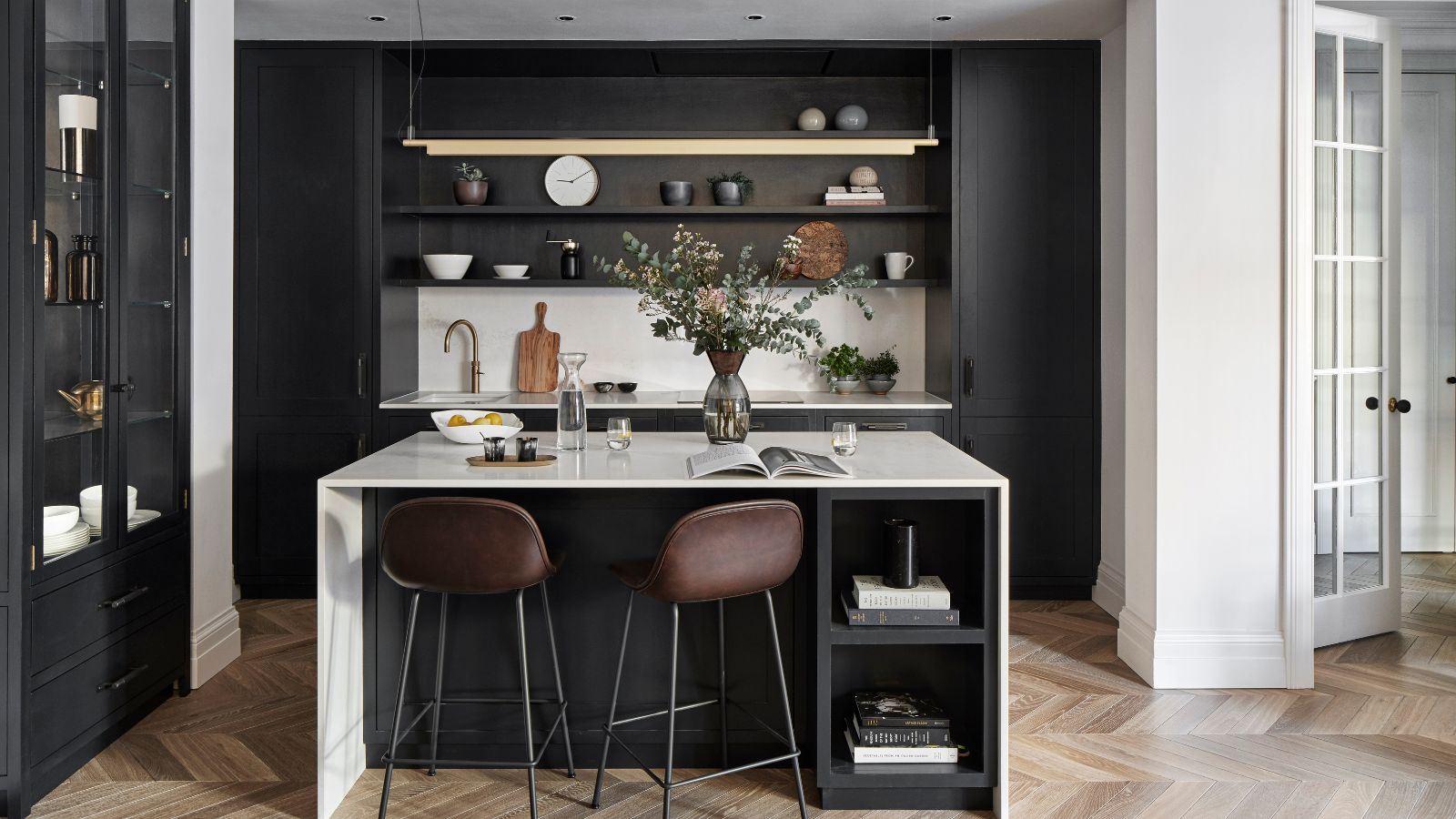 A black and white kitchen with a white marble island, brown leather bar stalls, brown parquet flooring, a white marble backsplash and shelving above.