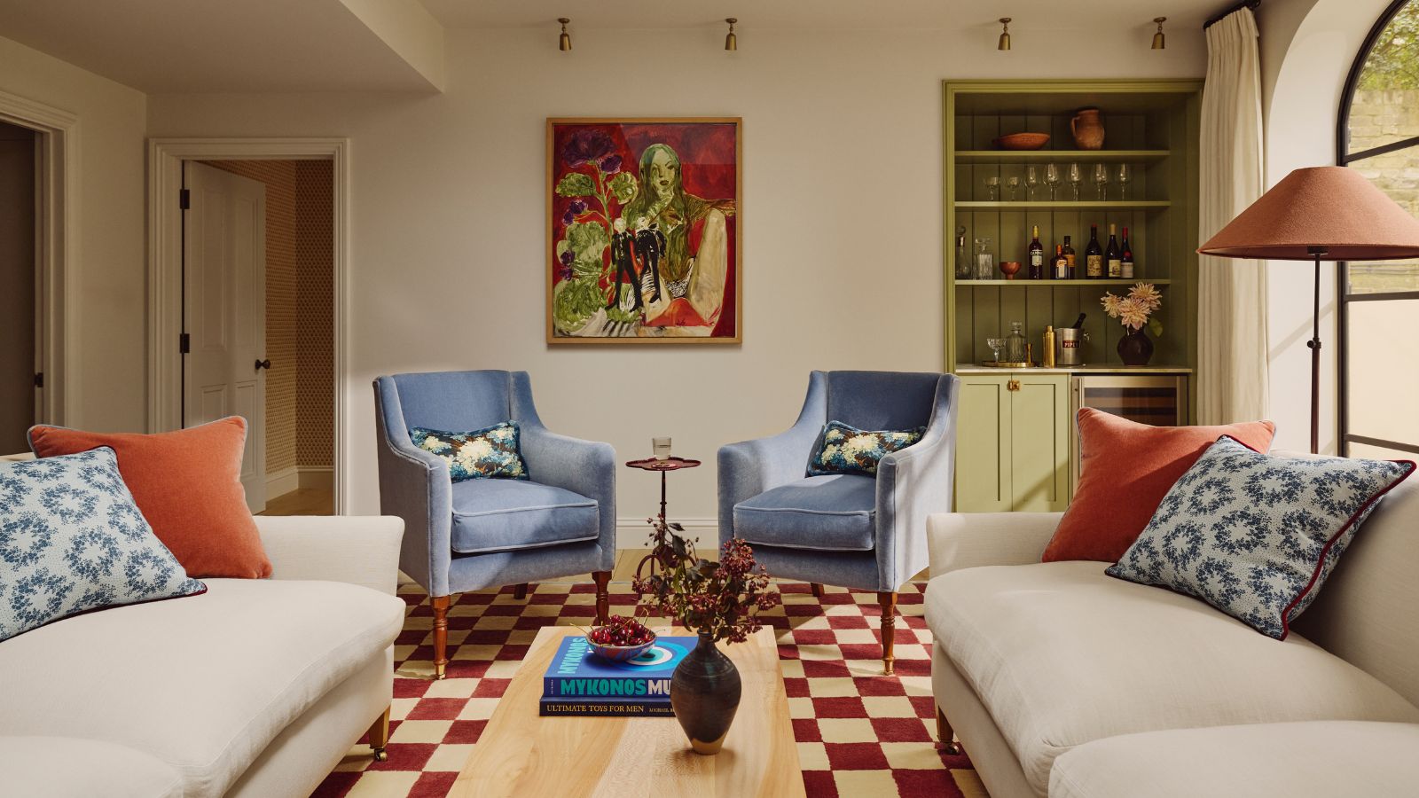 A maximalist living room with a red and white check tile floor, two cream sofas and two blue arm chairs, places symmetrically around a small wooden coffee table. 