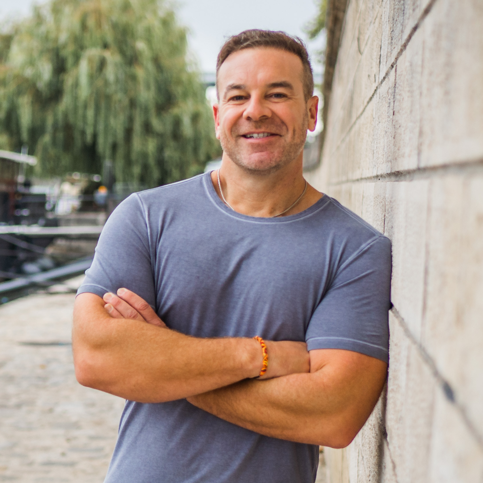 a photo of Bryan Clayton, CEO of GreenPal: a tanned man in a gray short sleeved shirt standing outside with his arms crossed facing the camera while leaning against a light gray brick wall