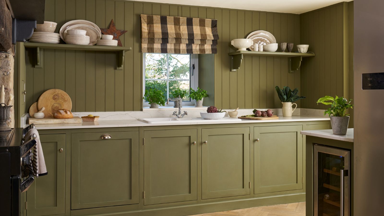 A small kitchen with matching painted green cabinets and shiplap walls. A plaid roman blind over a small window behind the sink. 