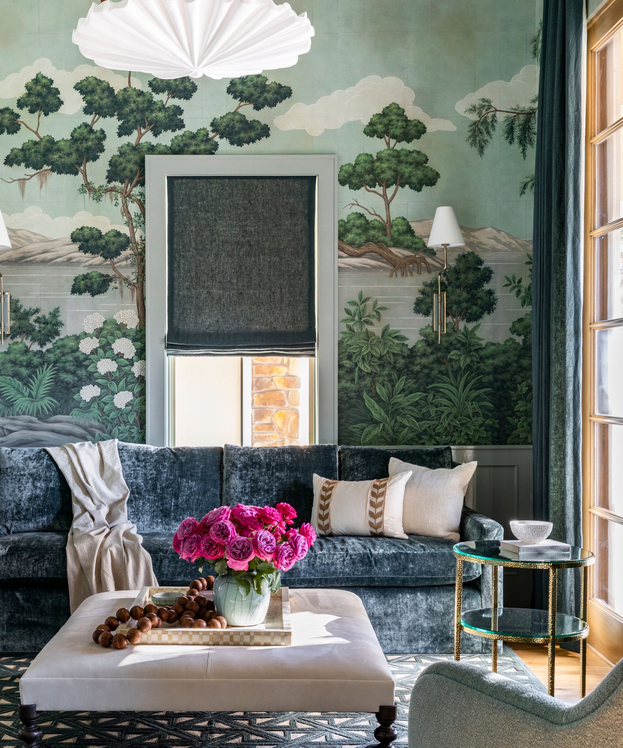 maximalist living room with wall mural, blue velvet. sofa and bright pink flowers on upholstered coffee table
