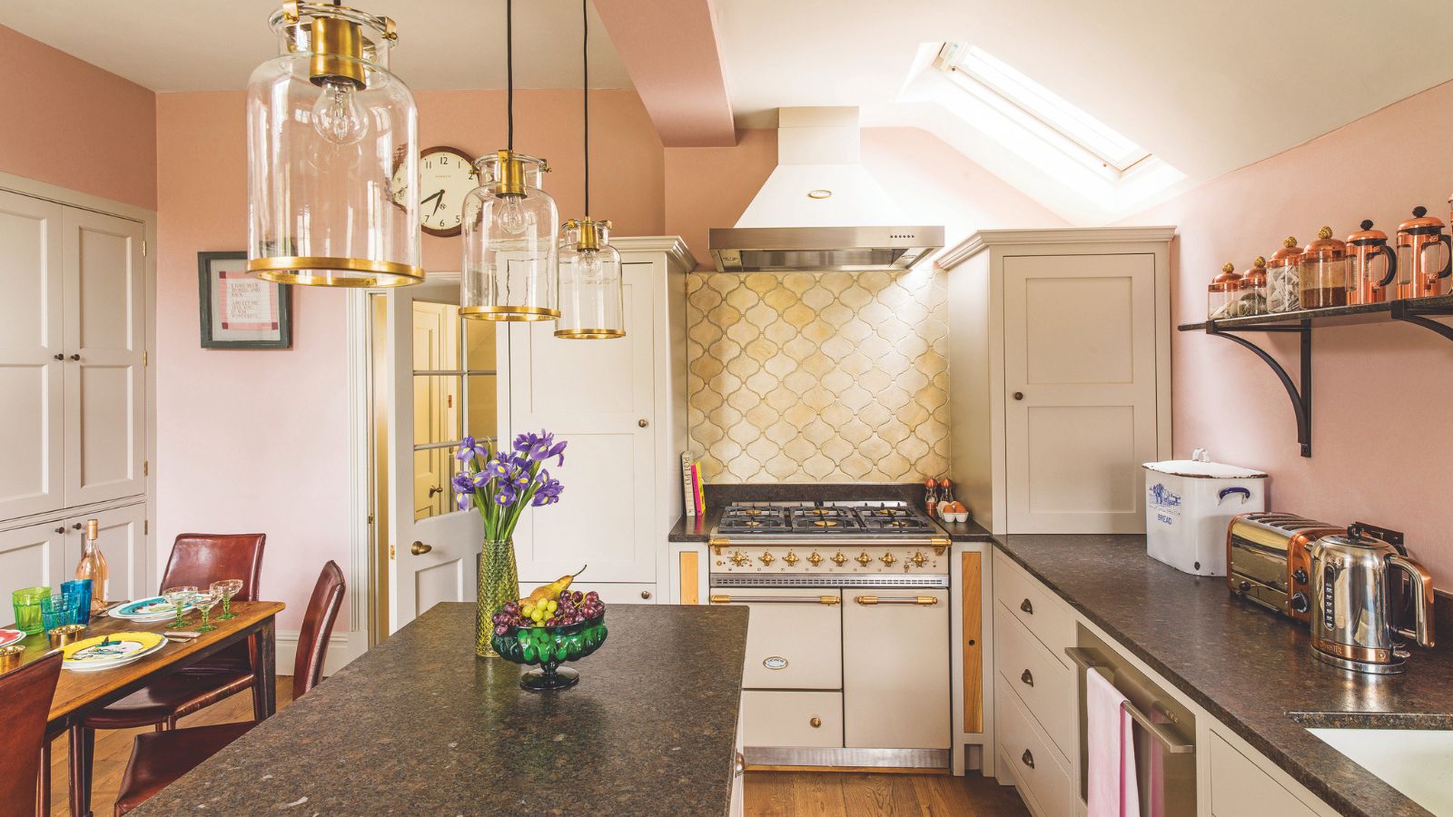 Kitsch pink kitchen with yellow shel-shaped backsplash tiles behind cream and gold range cooker. It&#039;s a kitchen diner with breakfast bar and gold accented pendant lights