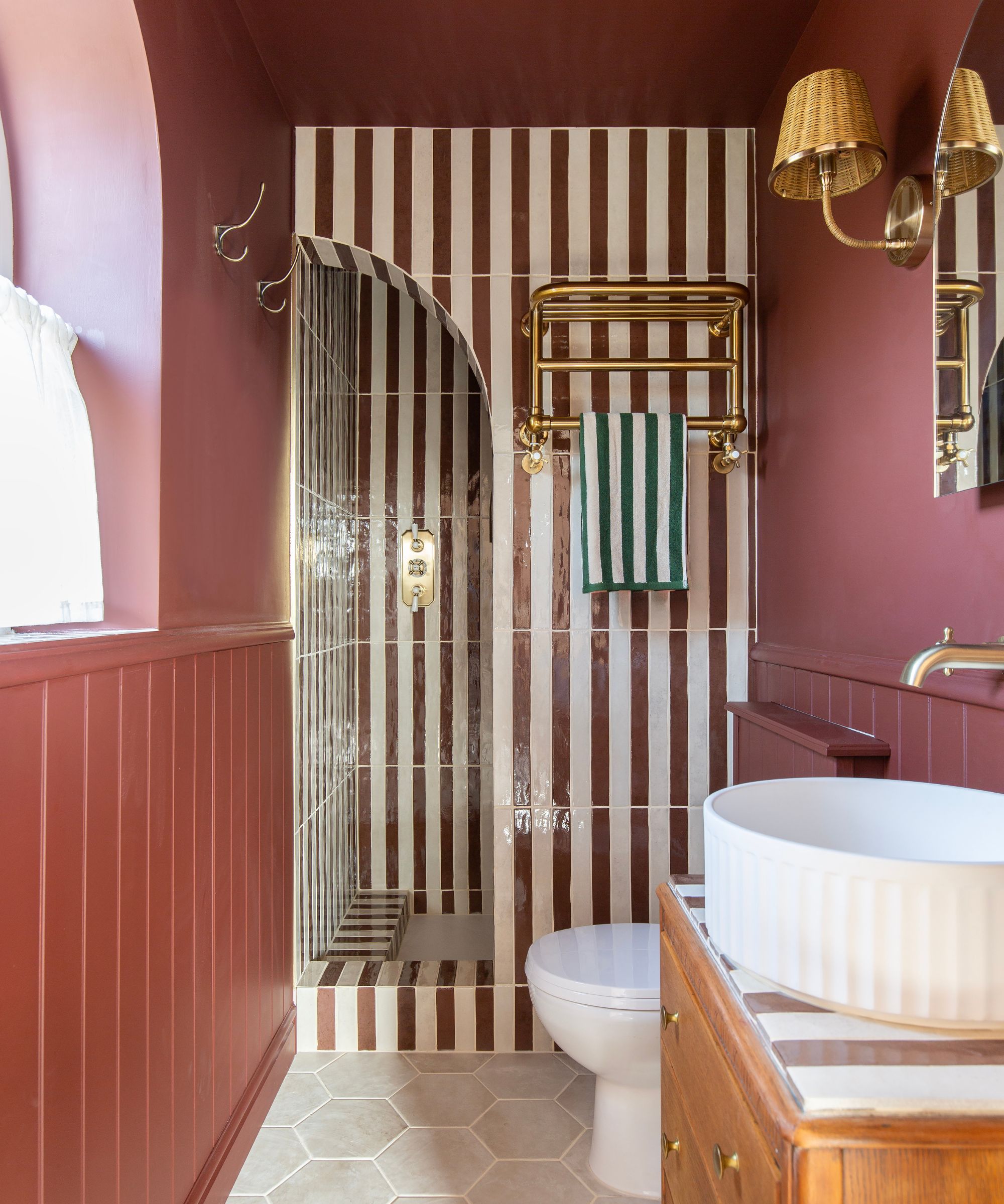 A long narrow bathroom with burgundy walls and a shower nook covered in red and white stripe tiles
